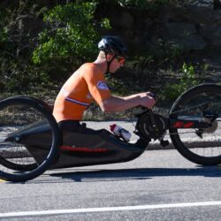 A man riding a hand cycle in orange Jersey at a high speed